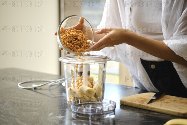 A person carefully adds sea buckthorn berries to a blender containing sliced bananas. The setting is a modern kitchen with natural light, highlighting the vibrant colors of the ingredients