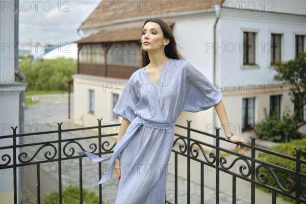 A woman stands on a balcony, wearing a flowing blue dress that drapes elegantly her slender body. The scenic backdrop features lush greenery and a charming building, under a soft cloudy sky