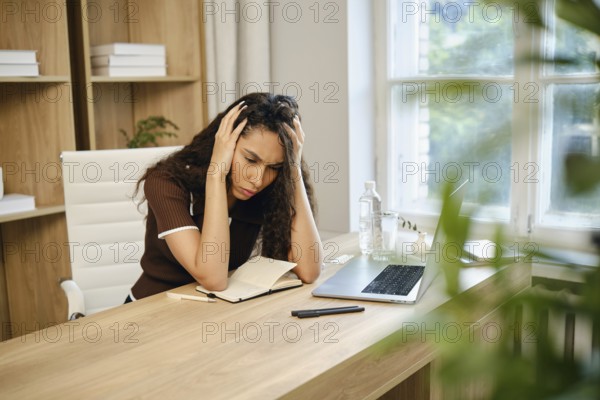 A young woman sits at her desk in a stylish home office, visibly stressed as she holds her head in her hands. A laptop, notebook, and water bottle are present, showing her work environment