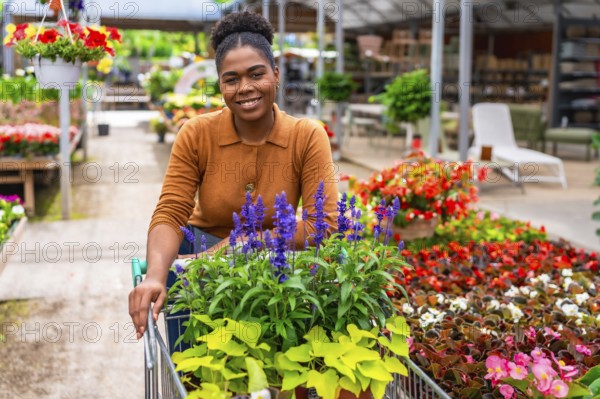 Smiling afro american woman pushing a shopping cart filled with vibrant flowers while browsing in a bustling garden center