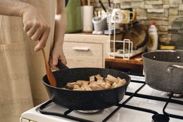 A person stirs diced pork meat in a frying pan on a gas stove. The kitchen is modern, featuring stone walls and organized utensils