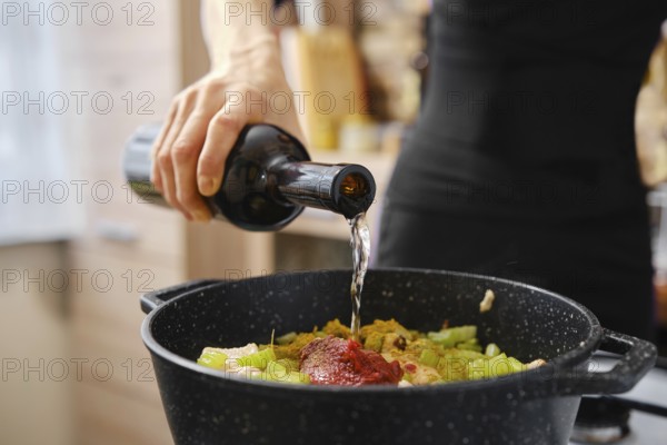 A hand pours liquid into a pot filled with chopped vegetables and sauce, creating a delicious mixture in a warm, inviting kitchen. This activity showcases culinary skills and creativity
