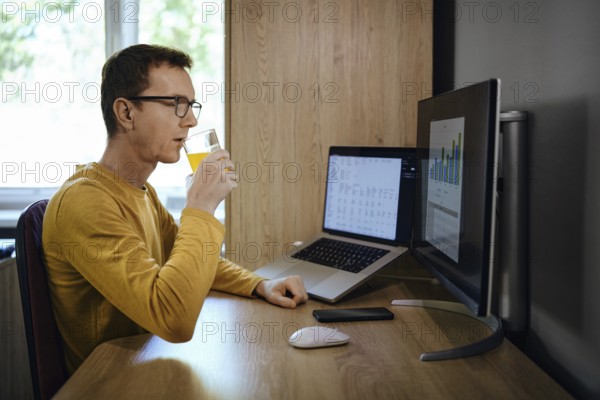 Individual is focused on work at a home office, sipping a drink while looking at two computer screens displaying charts. The workspace is organized with natural light coming through a window