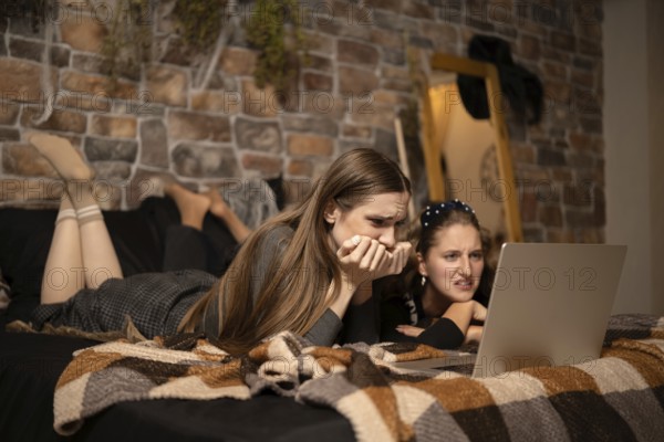 Two friends lie on a bed, deeply engaged as they watch an intense movie on a laptop. Their expressions show surprise and concern, set against a warm backdrop of brick walls and soft blankets
