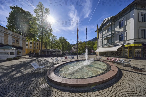 Wisbacherbrunnen, fountain, pedestrian zone, general architecture, cobblestones, public chairs, flagpoles, flags, trees, backlight of the sun, blue sky, cirrostratus clouds, intersection of Salzburger Straße with Wisbacher Straße and Ludwigstraße, Bad Reichenhall, district Berchtesgadener Land, Bavaria, Germany