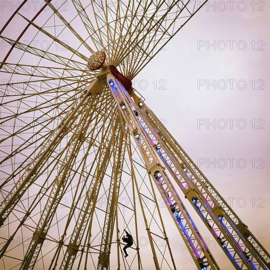 A maintenance worker is seen climbing the structure of a ferris wheel that features vibrant lights. The sky is a soft hue, indicating twilight as the worker ensures safety