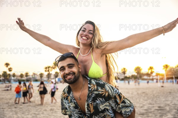 Carefree woman enjoying piggyback ride on man's back, spreading arms wide with joy on a sunny beach