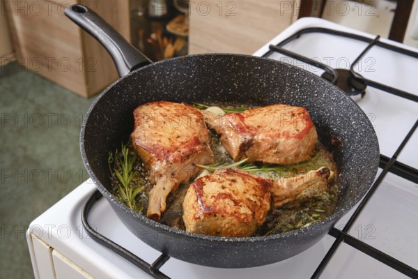Three pork rib roasts are sizzling in a skillet, infused with fresh herbs while cooking on a stovetop. The warm kitchen setting adds a cozy ambiance to this delicious meal preparation