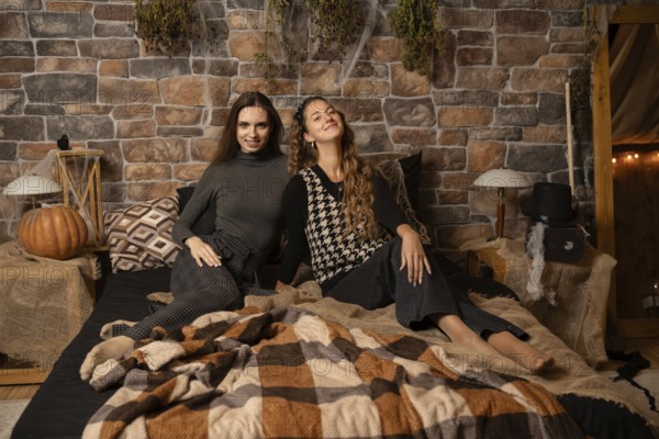 Two women relax together on a bed surrounded by autumn decor, including pumpkins and warm blankets, in a rustic setting adorned with stone walls and soft lighting