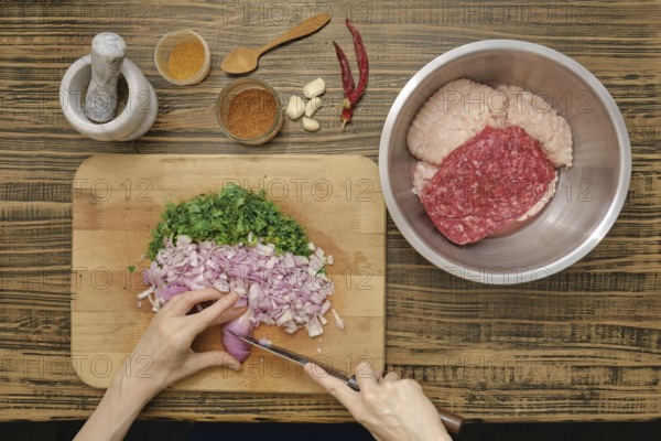 Hands chop shallots and parsley on a wooden cutting board. Ground meat is prepared in a bowl alongside spices and garlic. The kitchen setting is rustic with a mortar and pestle in view