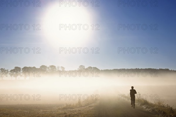 Man running on a dirt road in a rural landscape an autumn morning with low mist in backlight