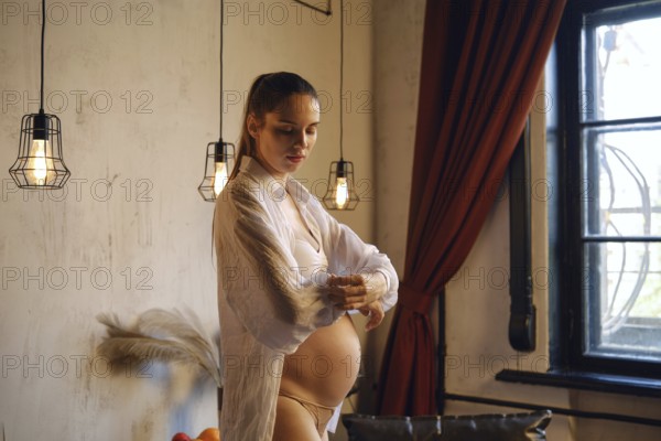 A pregnant woman stands in her elegantly decorated home, adjusting the sleeve of her shirt with a thoughtful expression