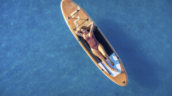 Lying on a paddle board, a woman enjoys the tranquility of the calm blue sea, soaking up the sun and embracing the peaceful summer vibes