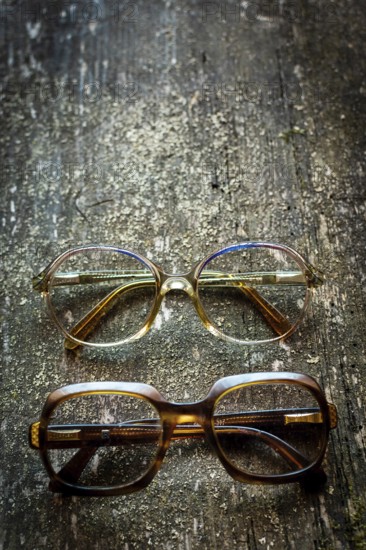 Two pairs of vintage eyeglasses rest on a weathered wooden surface, showcasing distinct styles. The top pair features a round frame, while the bottom displays a bold rectangular design