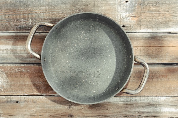 A gray frying pan with two sturdy handles on a weathered wooden tabletop. The pan is designed for even heat distribution through the thick bottom of the pan