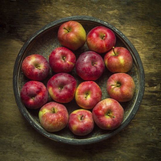 A collection of vibrant red apples fills a round bowl, placed on a textured wooden surface. The apples glisten, showcasing their freshness and inviting appearance