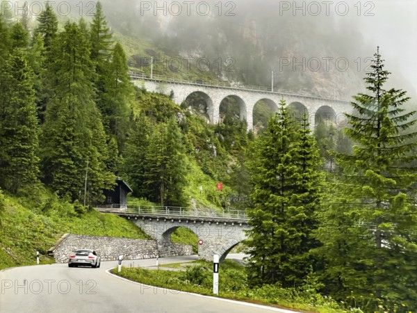 View of front Albula road Veja Alvra from Albula Pass towards Bergün with viaduct for railway in front Albulaviadukt I 1, above railway line to tunnel entrance, in the foreground below sports car Porsche GT3 on alpine road Bergstraße Passstraße, Bergün Bravuogn, Canton Graubünden, Switzerland