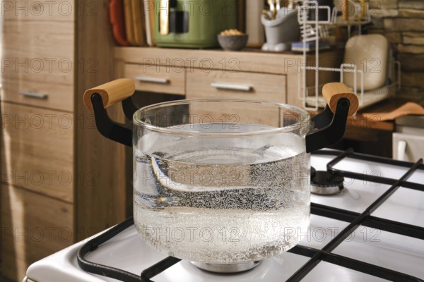 Water is boiling in a clear glass pot placed on a gas stove in a stylish kitchen. The scene showcases a clean, minimalist kitchen with natural wood tones and organized utensils