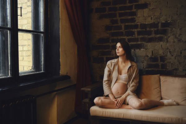A relaxed pregnant woman sits on a couch in her home, gazing thoughtfully out the window. Soft light filters through, highlighting her gentle pose and the warmth of the interior