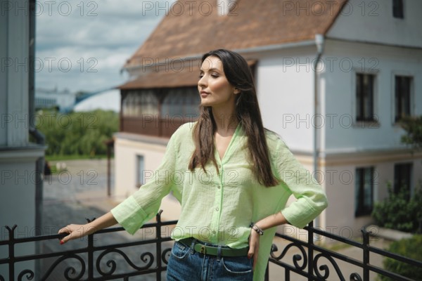 A young woman stands confidently on a balcony, wearing a light green blouse and blue jeans. She gazes thoughtfully into the distance, surrounded by a picturesque historic building under a bright sky