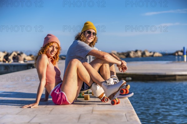 Smiling couple sitting on a sunny pier by the ocean, wearing casual attire and roller skates, embracing a carefree lifestyle