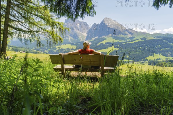 Person relaxing on a bench with a wide view of the mountains and green landscape, Alpe di Siusi, Dolomites, South Tyrol, Italy