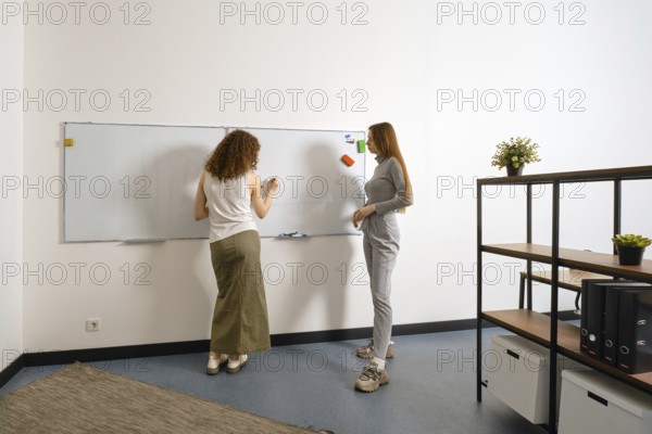 Two women collaborate on a whiteboard in a bright office dedicated to women's health. They discuss ideas and strategies of their startup
