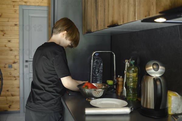 A boy washes vegetables in the sink and puts them in a colander, standing in profile at a sleek kitchen counter
