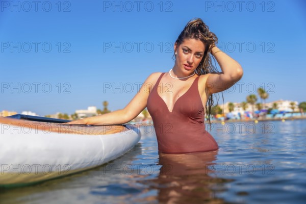 Young woman wearing swimsuit posing in the sea next to her paddle board, enjoying summer vacation on a sunny day