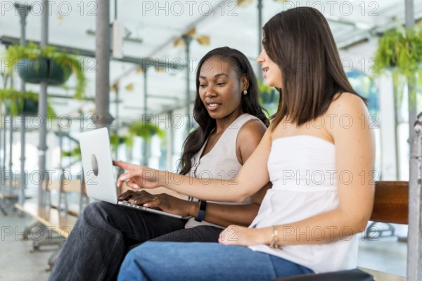 Two young businesswomen are sitting on a bench, using a laptop and discussing a project in a modern office space