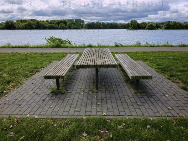 Empty picnic table with benches by a lake, surrounded by grass, under a cloudy sky in a park, summer, Eifel, Germany