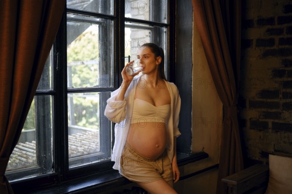 A pregnant woman stands by a large window at home, enjoying a drink from a glass. She wears a comfortable outfit and looks relaxed as natural light illuminates the cozy space