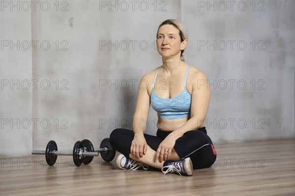 A woman sits cross-legged on a wooden gym floor, dressed in workout attire. Nearby, a pair of dumbbells rests on the floor, indicating a break after an exercise session