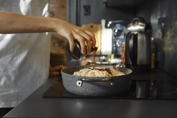 Unrecognizable person adds a soy sauce to a skillet filled with fresh vegetables and noodles on the stovetop