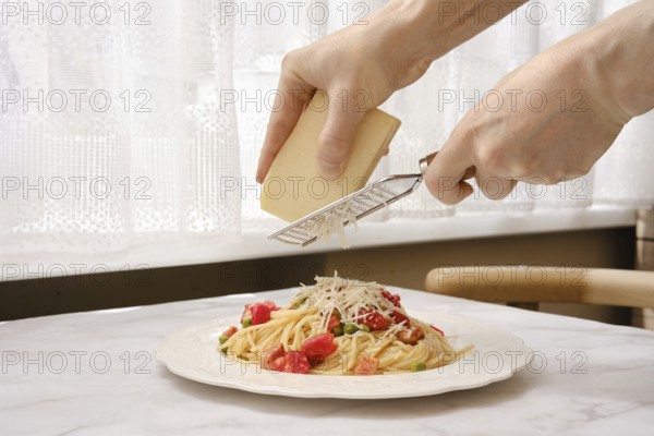 A person grates cheese onto a plate of spaghetti adorned with tomatoes and bacon in a well-lit kitchen. The cozy atmosphere is enhanced by the natural light filtering through sheer curtains