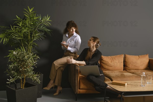 Two women engage in a lively discussion in a contemporary office. One holds a notepad while sitting on an armrest of a sofa, and the other sits with laptop on her knees