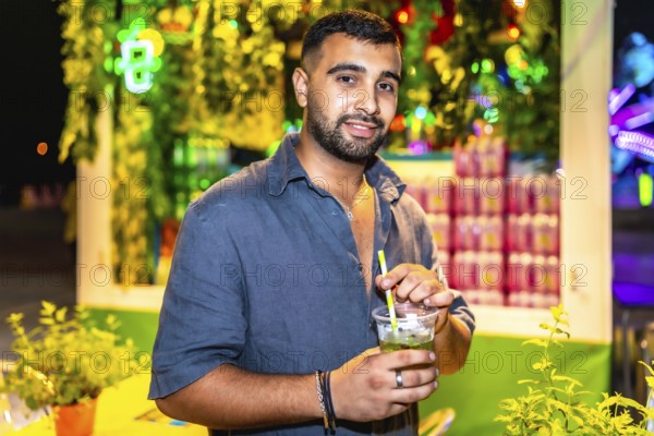 Stylish man savoring a vibrant mojito cocktail at a bustling night market, surrounded by colorful lights and greenery