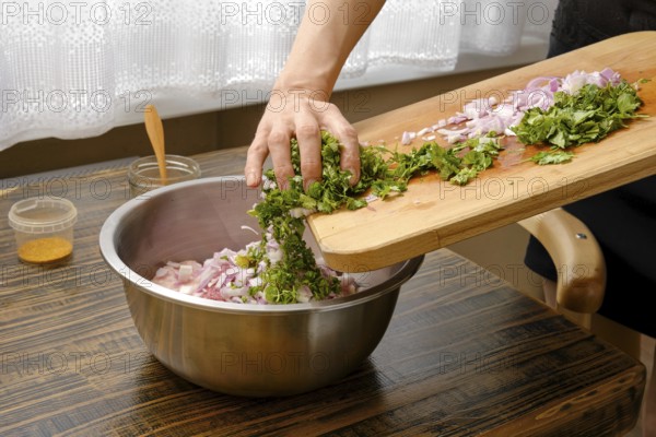 A hand adds chopped cilantro and onions into a silver mixing bowl with minced meat on a wooden table. The soft light from the window highlights the fresh ingredients