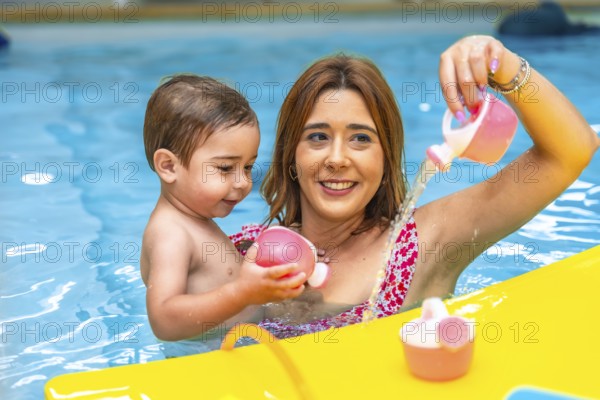 Happy mother and son having fun, playing with pink toys and pouring water in a swimming pool during summer vacation