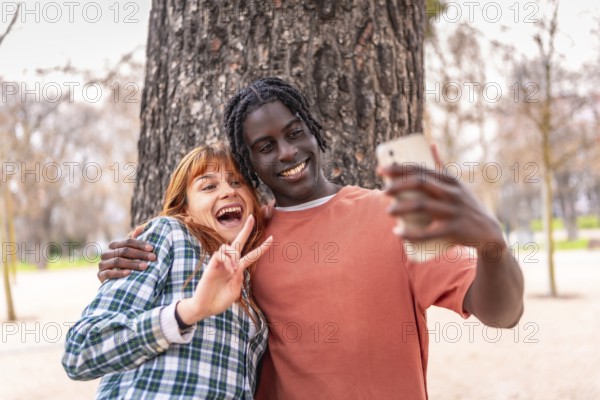 Two happy young students are making the victory sign while taking a selfie with a smartphone in a park