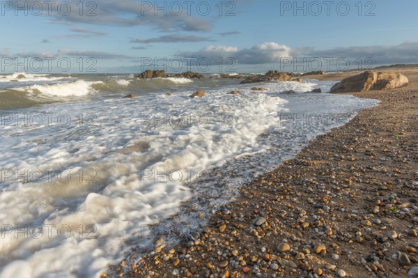 The waves crash gently against a rocky shoreline at sunset, creating a tranquil atmosphere. The sandy beach is strewn with pebbles and surrounded by a beautiful sky. Plage de l'Aubraie, Les Sables d'Olonne, Vendee, France