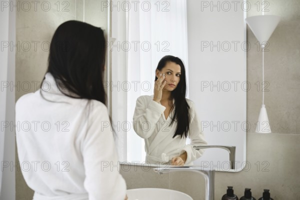 In a bright bathroom, a woman dressed in a white robe applies skincare to her face while looking into a mirror. Natural light fills the space, creating a serene atmosphere for self-care