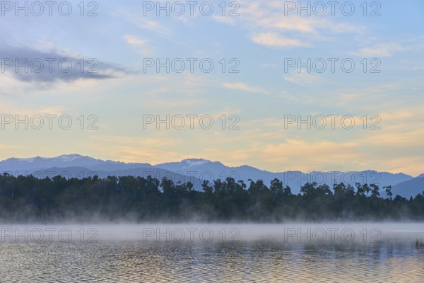 A peaceful lake with a misty surface and mountains in the morning light, Lake Mahinapua, Ruatapu, South Island, New Zealand