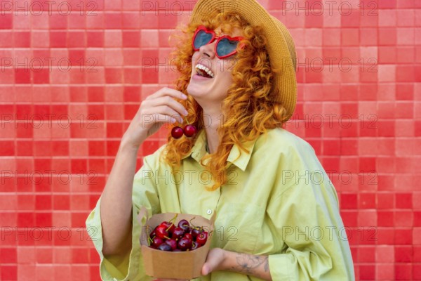 Young redhead woman with heart shaped sunglasses and straw hat is laughing while eating fresh cherries from a small cardboard box