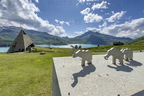 View from viewing platform to Mont-Cenis Pyramid Museum with stylised pyramid Museum in memory of Egypt campaigns of Emperor Napoleon Bonaparte and time in front of construction of reservoir, behind it Lac du Mont Cenis, in front on parapet small stylised figures of elephants Symbols for crossing of Alps Crossing of Alps by Hannibal, Mont Cenis Massif, Massif du Mont-Cenis, Graian Alps, Lanslebourg, Rhone-Alpes, France