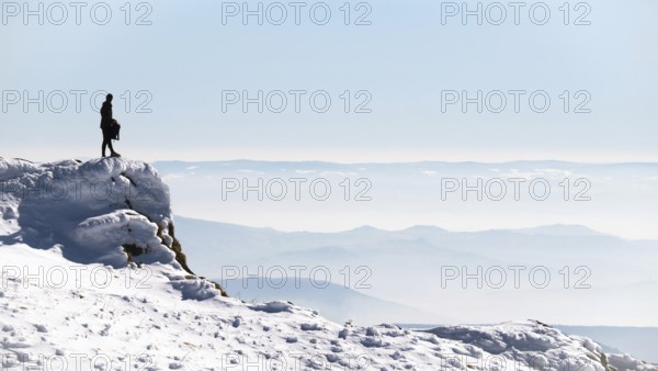 Standing on a rocky ledge, a walker takes in the breathtaking view of snow-covered mountains in Auvergne, France on a clear winter day. The expansive landscape stretches far into the distance