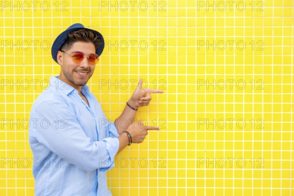 Fashionable man wearing a hat and sunglasses points to the side against a vibrant yellow tiled wall, presenting an ideal space for text or design elements