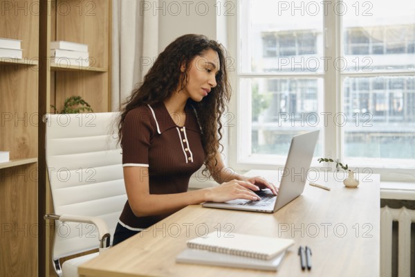 A woman with long curly hair is seated at a stylish wooden desk in a contemporary office. She is focused on typing on her laptop while surrounded by natural light streaming through large windows