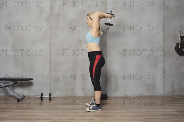 A focused woman in athletic wear is performing a tricep extension with a dumbbell in a well-equipped gym. The background features exercise equipment and an industrial design aesthetic