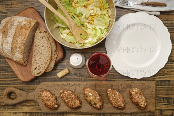A hearty meal is prepared with freshly made meat patties, a vibrant salad with corn and greens, and slices of rustic bread. A glass of red wine complements the meal, set on a wooden table